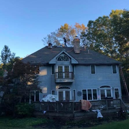 Two-story gray house with balcony, surrounded by trees and construction materials on the deck.