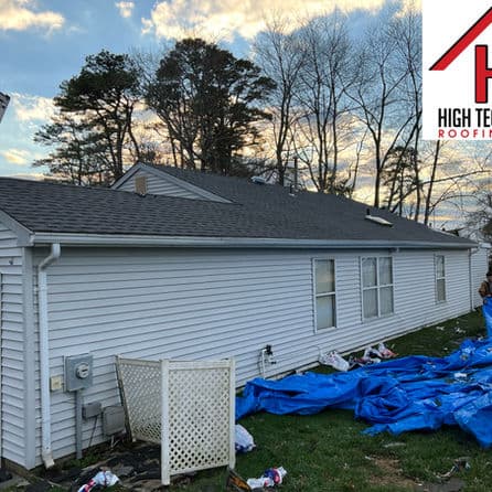 House with new roof installation, blue tarps on ground, trees and cloudy sky in background.