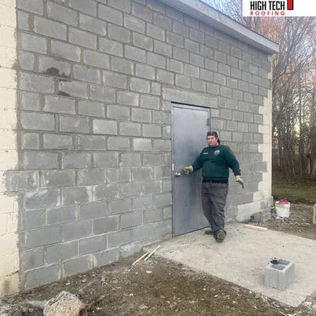 Man in green shirt standing by a newly built block wall and metal door for roofing project.