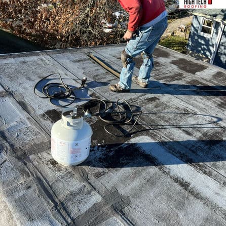 Person applying roofing material on a flat roof with tools and propane cylinder nearby.